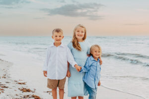 Three smiling children standing side by side on the beach at sunset, dressed in coordinated pastel clothing.
