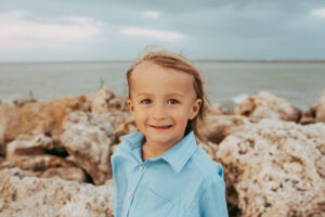 Young boy in a light blue button-up shirt smiling in front of rocky beach coastline.