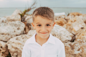 Young boy in a white dress shirt and khaki shorts standing barefoot on a beach with rocky shoreline.