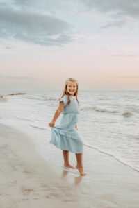 Smiling young girl in a light blue dress walking barefoot on the beach at sunset.