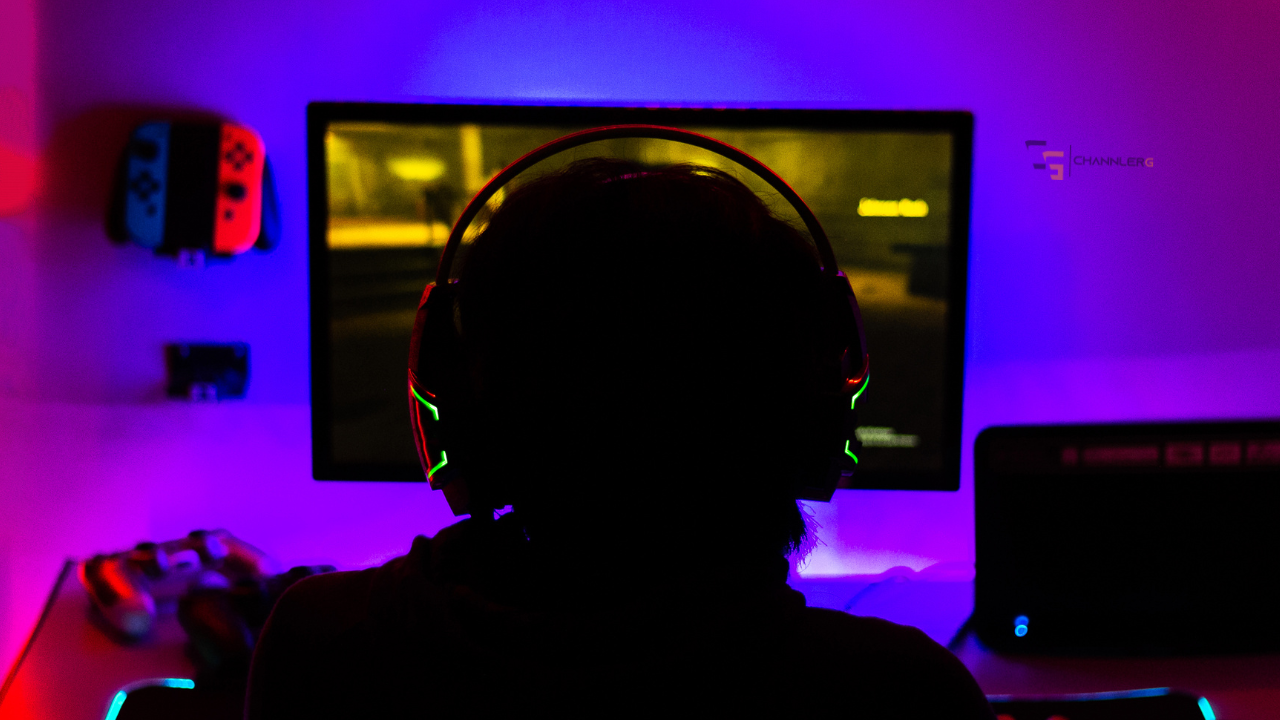 Back view of a person wearing a large gaming headset, silhouetted, facing a computer monitor in a room illuminated by purple and blue RGB lights.