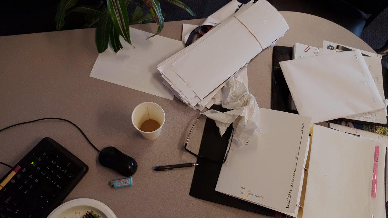 Cluttered office desk with paperwork, folders, coffee cup, keyboard, and office supplies.