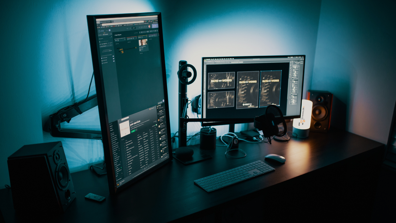 Dual monitor desk setup with vertical and horizontal screens, microphone, speakers, and ambient blue lighting.
