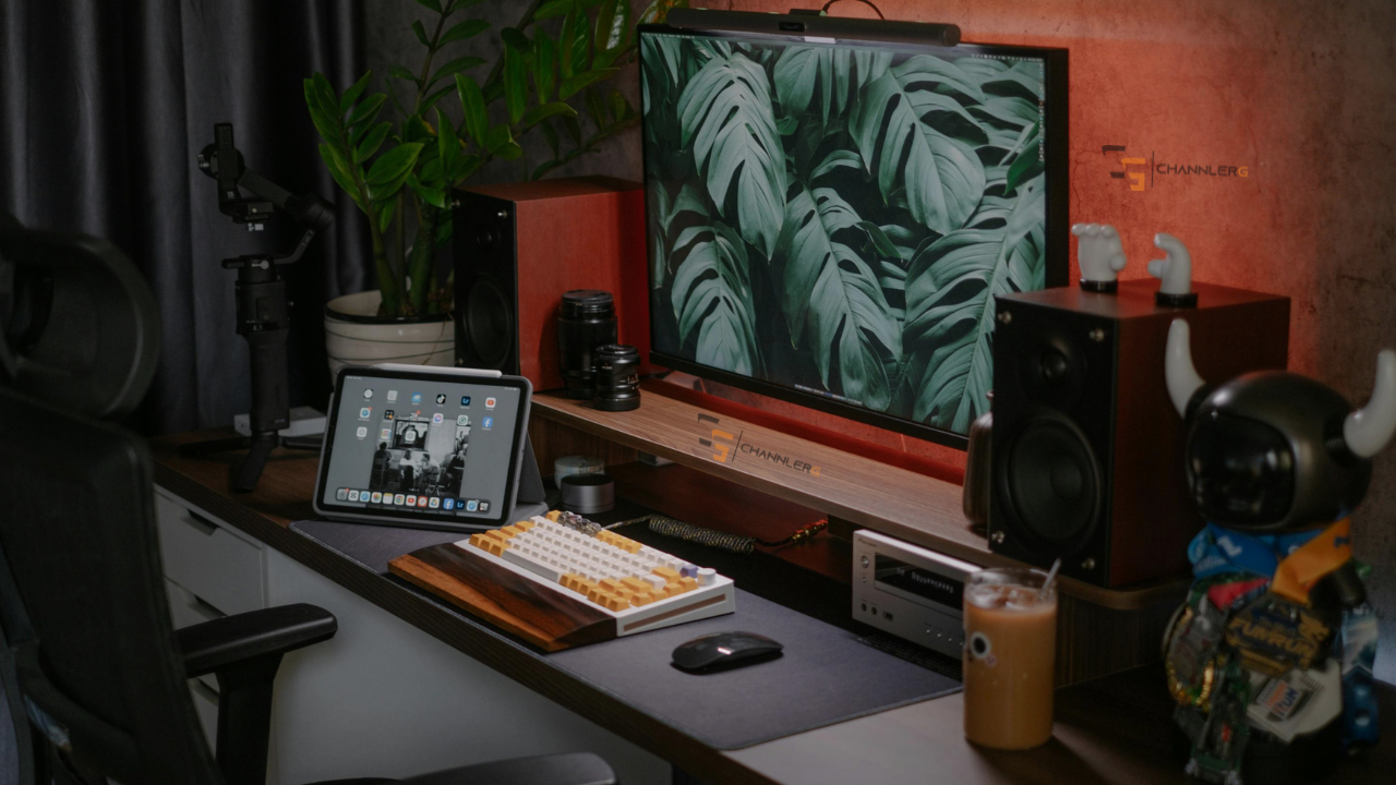 Modern desk setup with mechanical keyboard, monitor displaying green leaves, speakers, tablet, and warm ambient lighting.