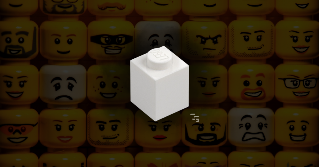 White LEGO brick displayed in front of a colorful wall of LEGO minifigure faces, representing gift ideas for adult LEGO fans.
